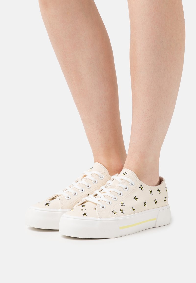Sneaker in tela beige con stampa floreale nera e gialla, suola in gomma bianca e lacci piatti. Presenta un design a taglio basso e accenti minimi.