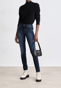 Suéter negro de punto, jeans ajustados azul oscuro y botas crema con cierre de cordones. El modelo sostiene un pequeño bolso negro con un logotipo colorido.