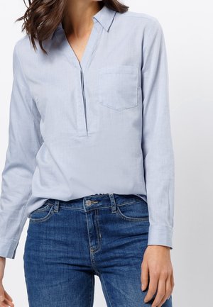 Vrouw die een lichtblauw overhemd met lange mouwen, een borstzak en blauwe spijkerbroek draagt, staand tegen een witte achtergrond.