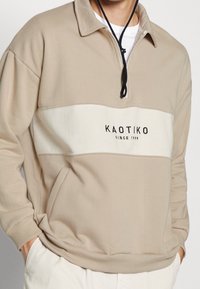 Beige hoodie i mjukt material, med ett krämfärgat horisontellt band och svart text. Dragkedja med snörning, ribbade ärmslut och nederkant.