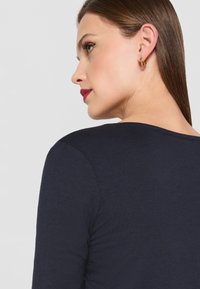 Námorně modré triko s dlouhým rukávem, zaobleným výstřihem a viditelným zadním pohledem; modelka má na sobě malé zlaté kruhové náušnice.