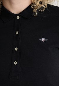Juodas polo marškinėliai, turintys faktūrinę medžiagą, klasikinį apykaklę ir penkių mygtukų užsegimą. Mažas baltas išsiuvinėtas logotipas ant krūtinės.