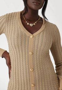 Cardigan beige a maglia con scollo a V, cinque bottoni crema e polsini a coste. Gli accessori includono una collana a forma di cuore in tono oro.