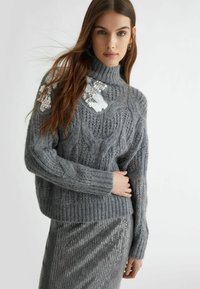 Maglione grigio a maglia con collo alto, caratterizzato da dettagli di paillettes sulle spalle. Abbinato a una gonna argentata con paillettes.
