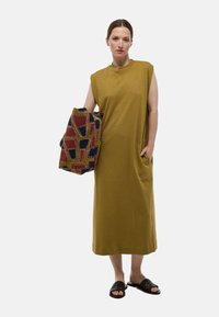 Robe sans manches vert olive avec une coupe ample et des poches latérales, accompagnée d'un châle à motifs comportant des formes rouges, bleues et jaunes.