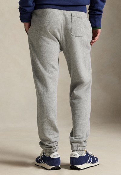 Persona che indossa pantaloni della tuta grigi con polsino elastico alla caviglia e sneakers blu navy, in piedi con le mani dietro.