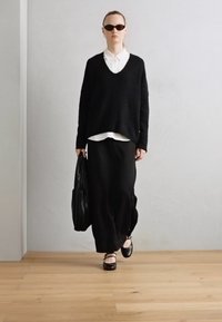 Maglione nero oversize con scollatura a V, indossato sopra una camicia bianca con colletto. Gonna nera lunga, scarpe ballerine scure e una grande borsa a tracolla nera con texture.