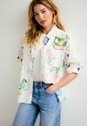 Young woman with long blonde hair wearing a white button-up shirt with colorful fruit and floral prints, paired with light blue jeans.