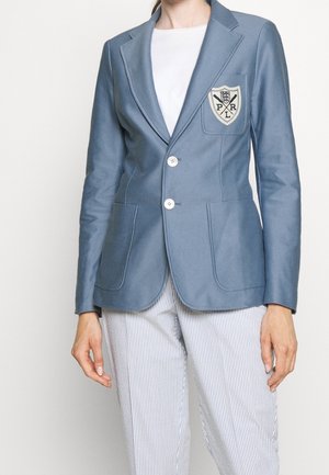 Kobieta ubrana w niebieską marynarkę z dwoma białymi guziczkami i naszywką na lewej piersi, białą koszulę oraz spodnie w pionowe paski.