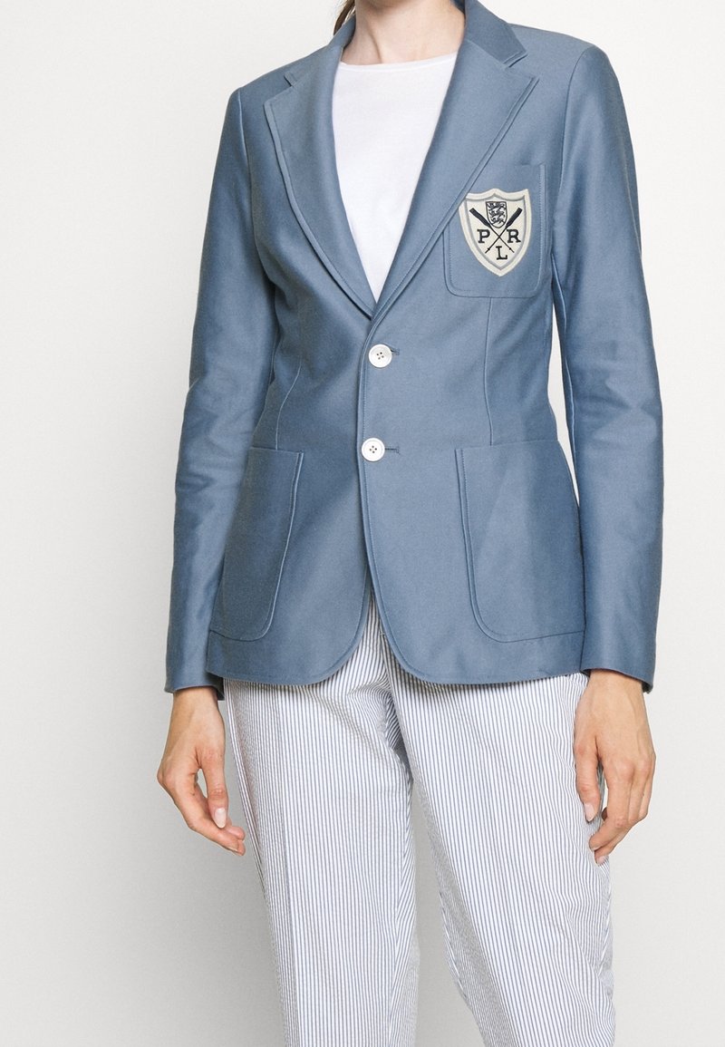 Femme portant un blazer bleu avec deux boutons blancs et un écusson sur la poitrine gauche, une chemise blanche et un pantalon à rayures verticales.