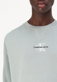 Maglione a maglia verde chiaro con colletto rotondo a coste, che presenta un piccolo logo bianco "CK" e il testo "Calvin Klein Jeans" in nero.