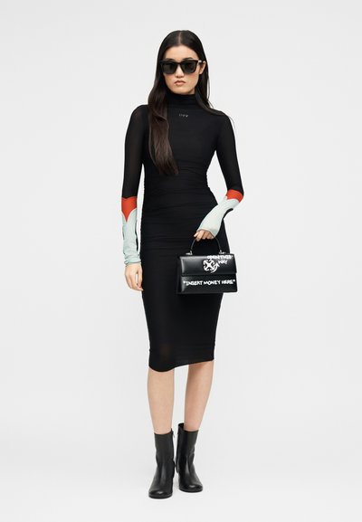 Γυναίκα που φοράει στενό μαύρο φόρεμα με κόκκινες και λευκές λεπτομέρειες στα μανίκια, μαύρα γυαλιά ηλίου, μαύρες μπότες, κρατώντας μια μαύρη τσάντα με λευκό κείμενο.