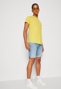 Camiseta amarilla de manga corta combinada con pantalones cortos de denim azul claro, zapatillas blancas y gafas de sol amarillas de gran tamaño, exhibidas sobre un fondo liso.