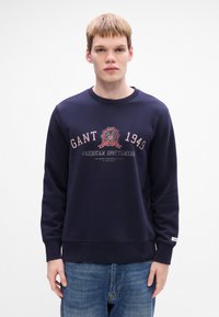 Sötétkék pulóver kerek nyakkal, hosszú ujjakkal, és nyomtatott logóval, amelyen a "GANT 1949" és az "American Sportswear" feliratok találhatóak.