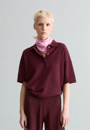 Person mit kurzen blonden Haaren trägt ein bordeauxrotes, strukturiertes Kurzarmhemd und eine Hose, dazu einen rosa Seidenschal um den Hals gebunden.