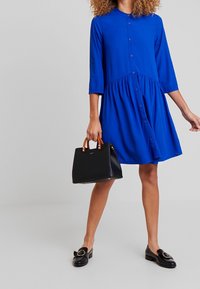 Robe bleue boutonnée avec col et manches trois-quarts, dotée d'une taille froncée et d'une silhouette fluide. Associée à un sac à main noir et des mocassins.