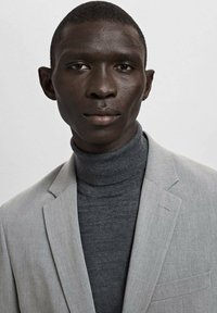 Jonge man met een donkere huid, gekleed in een grijze coltrui en een lichtgrijze blazer, kijkt recht in de camera tegen een effen achtergrond.
