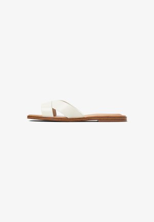 Sandal flat con amplias correas de cuero blanco entrelazadas y suela de madera marrón, vista de perfil sobre un fondo blanco.