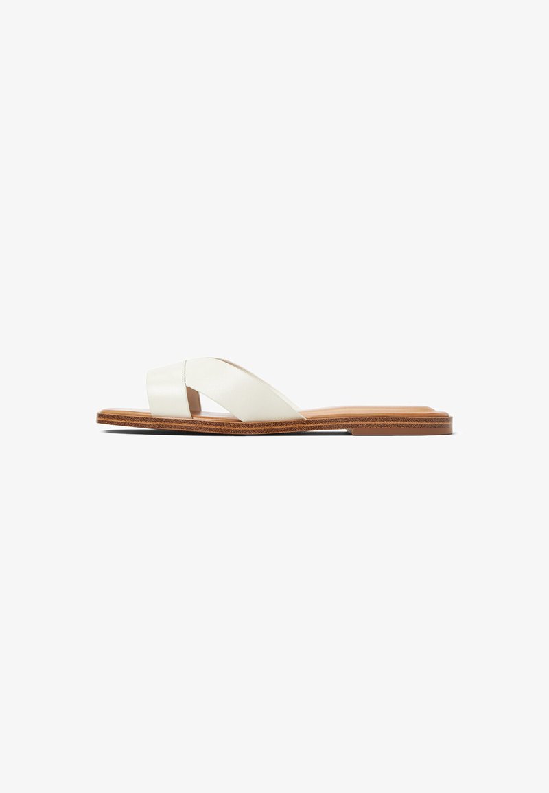 Sandale plate avec de larges brides en cuir blanc entrecroisées et une semelle en bois brun, vue de côté sur fond blanc.