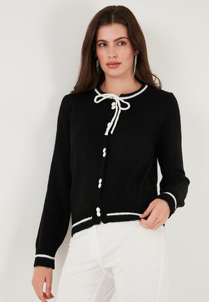 Cardigan nero con bordi bianchi, dotato di scollatura rotonda, laccetto e chiusure a bottoni. Maniche lunghe con polsini a contrasto.