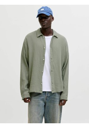 Mann mit blauer Kappe, hellgrünem Hemd mit Knöpfen über einem weißen T-Shirt und locker sitzender, ausgewaschener blauer Jeans, der vor einem einfachen Hintergrund steht.