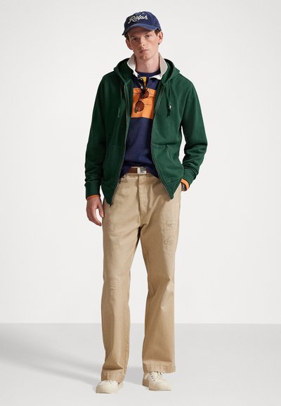 Jovem vestido com uma camisa azul e laranja sob um hoodie verde com fecho, calças bege, ténis brancos e um boné azul com o texto "Ralph".