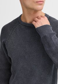 Pull en tricot gris foncé avec un motif texturé. Présente un col et des poignets côtelés, mettant en avant un style de manches raglan ajustées.