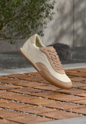 Kremowo-beżowy sneaker z teksturowaną cholewką, sznurowadłami i gumową podeszwą, prezentowany na tle drewnianej powierzchni z kamykami.