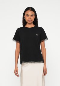 Črna majica s tilskimi dodatki na rokavih in spodnjem robu. Na prsih ima majhen zlati okrasni detajl. Kratki rokavi in sproščen kroj.