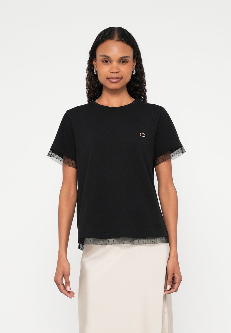 Črna majica s tilskimi dodatki na rokavih in spodnjem robu. Na prsih ima majhen zlati okrasni detajl. Kratki rokavi in sproščen kroj.