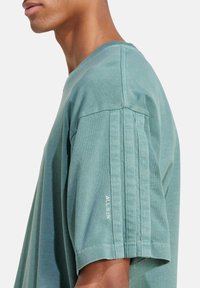 Zelené bavlněné tričko s volným střihem, krátkými rukávy a vertikálním pruhovaným detailem na ramenech. Na rukávu je nápis "ALL SZN".