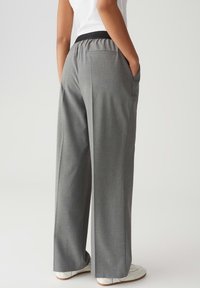 Persona che indossa pantaloni larghi grigi con cintura elastica nera e sneakers bianche, con le mani in tasca, in piedi contro uno sfondo semplice.