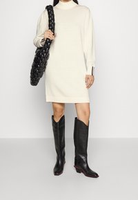 Femme portant une robe pull crème, des bottes noires jusqu'aux genoux avec des bordures roses, et tenant un grand sac à main en cuir tressé noir.
