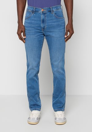 Uomo con jeans blu a gamba dritta e scarpe da ginnastica bianche, in piedi davanti a uno sfondo chiaro e semplice.