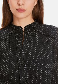 Schwarze Bluse mit kleinem weißen Punktmuster, Schlüsselhalsschnitt mit Knopfverschluss und Rüschenschultern. Leichtes Material mit strukturierter Oberfläche.