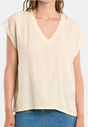 Donna che indossa un top beige largo, senza maniche e con scollatura a V, abbinato a jeans di denim blu.