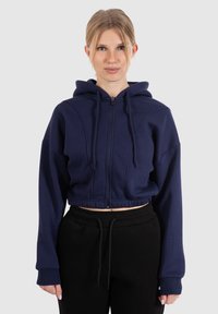 Felpa cropped blu navy in morbido tessuto, con zip frontale, cappuccio con cordino e polsini a costine. Abbinata a pantaloni della tuta neri.