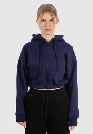 Haut en molleton bleu marine coupé, fabriqué dans un tissu doux, avec une fermeture éclair à l'avant, une capuche à cordon et des poignets côtelés. Assorti à un pantalon de jogging noir.