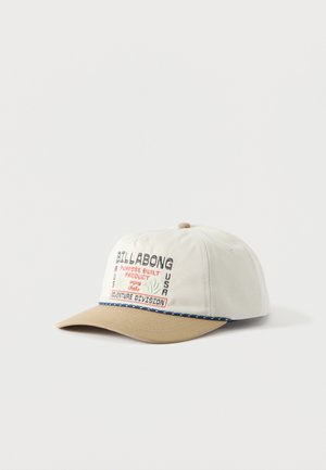 Gorra Billabong blanca y beige con texto y logo bordados, con la inscripción "Adventure Division" y costura decorativa azul en el borde de la visera.