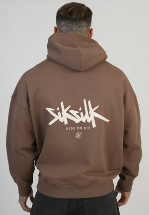 Mężczyzna z krótkimi włosami i tatuażem na szyi, ubrany w brązową bluzę z kapturem z białym napisem „SikSilk Ride or Die” na plecach, stojący na jednolitym tle.
