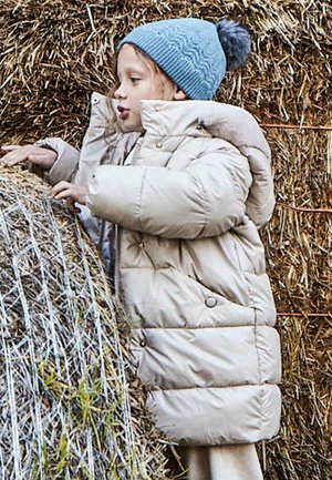 Light beige puffer jacket with a high collar and snap buttons. Blue knitted beanie with a pom-pom atop, against a backdrop of hay bales.