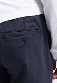 Marineblauwe broek met een gestructureerde afwerking, voorzien van een achterzak, knoopsluiting en een klein merklabel.