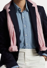 Marinblå jacka över en ljus denimskjorta, med en ljusrosa kabelstickad tröja draperad över axlarna. Vita byxor fulländar outfiten.