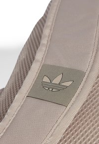 Primer plano de una correa de mochila beige con bordes cosidos y un parche del logo Trefoil de Adidas.