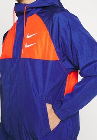 Námořnická modrá a oranžová větrovka s kapucí, elastickými manžetami, bočními kapsami a výrazným bílým logem Nike na hrudi. Hladká textura.