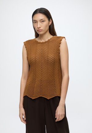 Femme portant un top marron sans manches en crochet avec des bords festonnés et un pantalon ample foncé, debout devant un fond blanc uni.