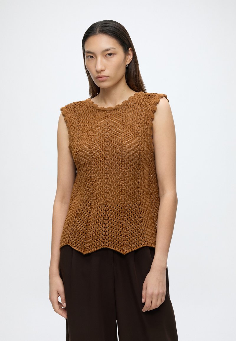 Femme portant un top marron sans manches en crochet avec des bords festonnés et un pantalon ample foncé, debout devant un fond blanc uni.