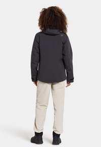 Veste noire imperméable avec capuche, pantalon beige et bottes noires. La veste présente une texture lisse et un logo sur le haut du dos.