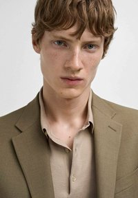 Brauner Blazer über einem beigen Poloshirt mit strukturiertem Finish. Kurze, wellige Haare und helle Haut mit Sommersprossen. Neutraler Hintergrund.