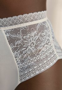 Chiloți albi cu talie înaltă, din dantelă, cu modele florale, margine delicată de-a lungul taliei și textură canelată pe laterale.